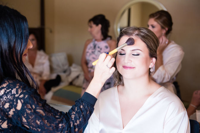 Bride in a satin robe smiling with eyes closed as a makeup artist applies powder with a large brush, bridesmaids and a mirror visible in the bridal suite during wedding morning preparations.