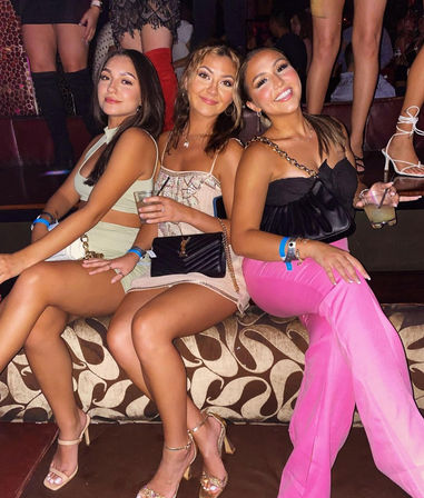 Three friends smiling in a nightclub lounge booth, dressed up in party outfits and heels, holding cocktails for a fun night out.