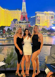 Three women in cocktail dresses posing on a balcony over a fountain lake at night with the illuminated Las Vegas Strip skyline and Eiffel Tower replica in the background.