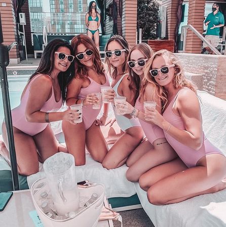 Five friends in matching pastel swimsuits and sunglasses toasting with drinks on a rooftop hotel pool daybed, ice bucket and pitcher in the foreground and city buildings in the background — playful rooftop pool party vibe.