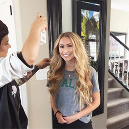 Smiling young woman with long wavy blonde hair getting a final spritz from a stylist in a cozy home hallway by a mirror and staircase.