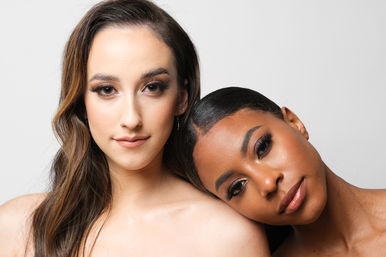 Close-up studio beauty portrait of two women with polished makeup, one leaning her head on the other’s shoulder against a neutral background.