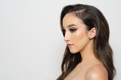 Glam profile studio portrait of a woman with long dark hair, bold winged eyeliner, luminous cheek highlight, and multiple ear piercings on a plain white background.