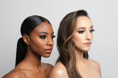 Beauty studio portrait of two women with glam makeup and neutral gray background — one with a sleek low ponytail and glowing skin, the other with long wavy hair and soft contouring.
