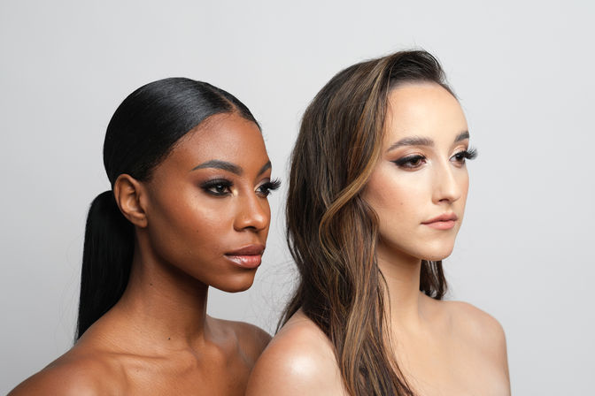 Beauty studio portrait of two women with glam makeup and neutral gray background — one with a sleek low ponytail and glowing skin, the other with long wavy hair and soft contouring.