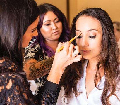 Bridal makeup session: artist applying eyeshadow to a woman with long hair and false lashes while a tattooed assistant watches in a warm getting‑ready room.