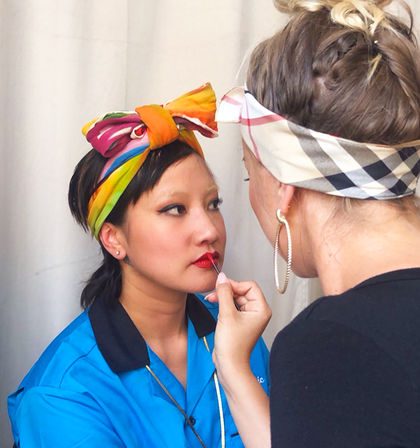 Close-up of a makeup artist using a brush to apply bold red lipstick to a seated woman wearing a colorful rainbow headscarf and blue shirt in a beauty studio.