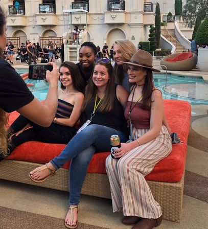 Five young women smiling and posing on an orange poolside lounge at a hotel resort pool while a friend takes their photo on a smartphone, casual summer outfits and relaxed outdoor pool deck vibe.