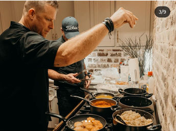 Two cooks seasoning food over a busy home kitchen stovetop with multiple pots and pans—fried patties, simmering sauces and a pot of rice against a brick backsplash, casual meal prep vibe