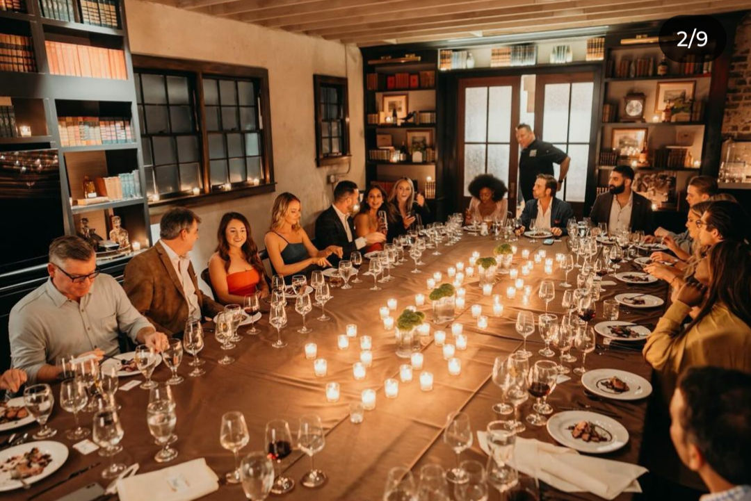 Cozy candlelit private dining room with a long table set for a formal group dinner, adults in evening attire sharing wine and plated courses amid bookshelves and warm ambient lighting.