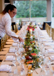 Server arranging glassware on an elegant, candlelit long banquet table with white linens, gold chairs, greenery, grapefruit halves and floral centerpieces on a covered patio event.