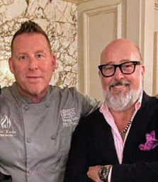 Smiling chef in a gray coat and a well-dressed guest in a blazer with a pink shirt and pocket square posing together in an upscale restaurant interior with marble wall