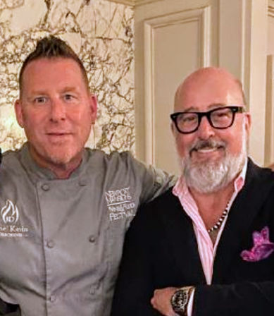 Smiling chef in a gray coat and a well-dressed guest in a blazer with a pink shirt and pocket square posing together in an upscale restaurant interior with marble wall