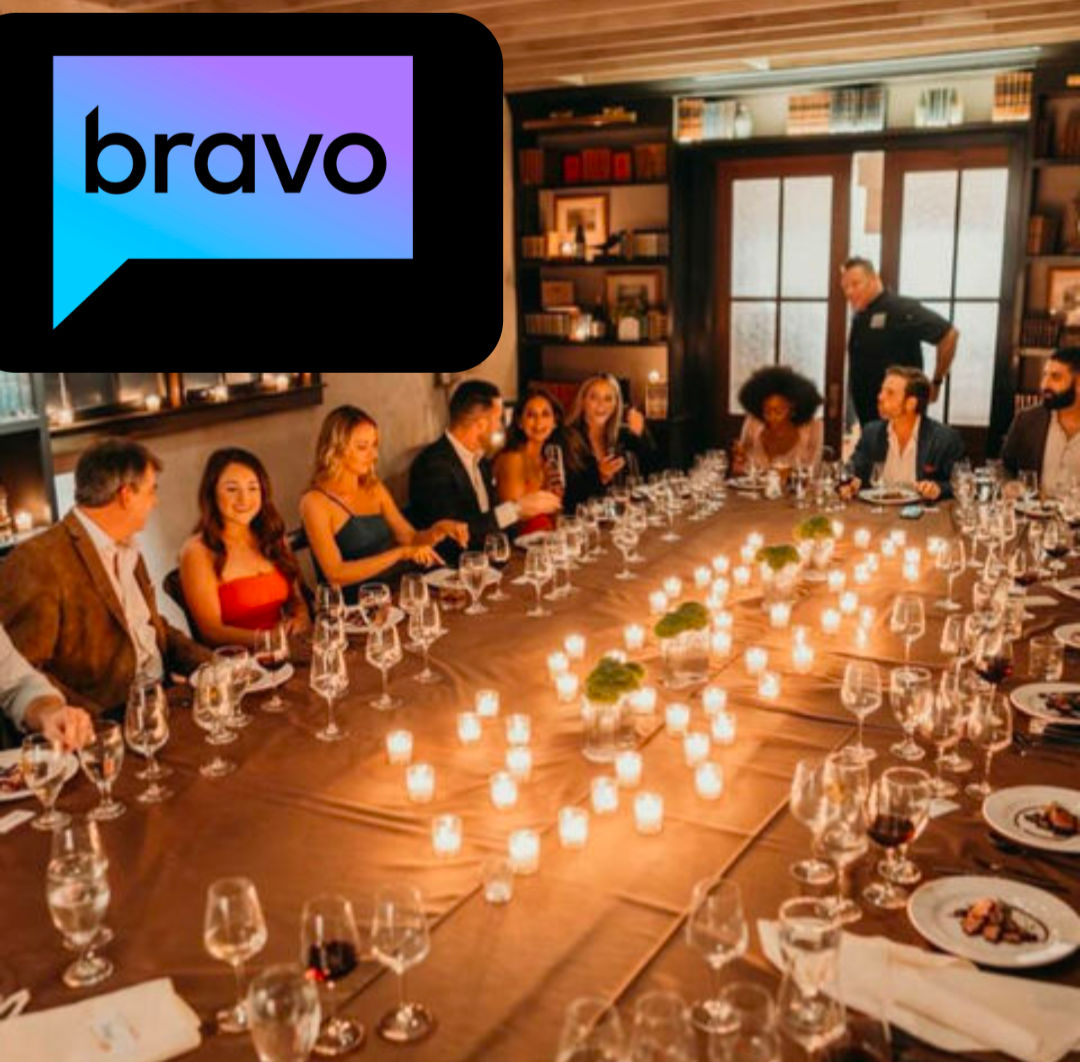 Upscale candlelit long-table dinner in a private dining room with a group of adults, many wine glasses and plated appetizers, votive candles down the center and a colorful speech-bubble logo in the top-left corner.