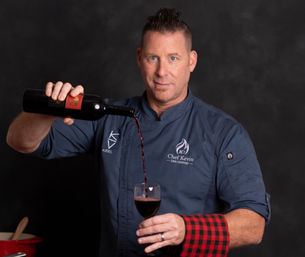 Chef portrait pouring red wine into a glass, man in navy chef coat with a red-and-black checkered towel on his wrist against a dark studio backdrop.