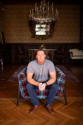 Indoor portrait of a man in a gray t-shirt and blue pants sitting on a red-and-green plaid armchair in an ornate parlor with a crystal chandelier, gilded mirror, antique furniture and hardwood parquet floor.