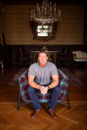 Casual portrait of a man seated on a red-and-green plaid armchair in an ornate historic mansion parlor with a crystal chandelier, gilded mirror, and parquet floor.