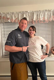 Smiling man in a chef jacket and woman wearing a “Bride” t‑shirt pose together indoors in front of window blinds and a rose-gold bridal shower banner.