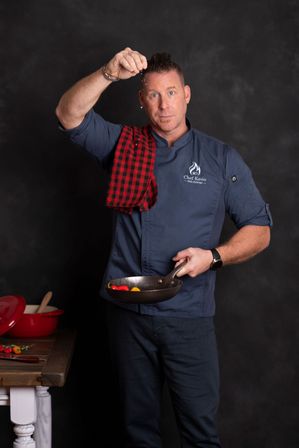 Studio portrait of a chef in a navy jacket with a red checkered towel, sprinkling salt into a frying pan with colorful mini peppers, dark backdrop and wooden table with a red Dutch oven.