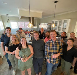 Cheerful group of friends gathered in a modern open-concept home kitchen, posing for a casual house party photo under hanging pendant lights, with some people holding drinks and smiling at the camera.