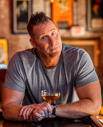 Man in a gray t-shirt leaning on a bar counter, holding a coupe glass of amber cocktail in a warm-lit, exposed-brick pub with framed posters