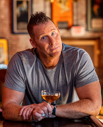 Muscular man in a gray t-shirt leaning on a bar counter, holding a coupe glass with an amber cocktail and giving a quizzical smile in a warm, brick-walled cocktail bar with blurred framed posters