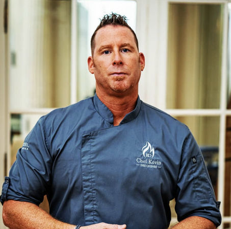Restaurant chef portrait: man in slate-gray chef coat with flame logo standing indoors in front of glass doors, arms folded and neutral expression