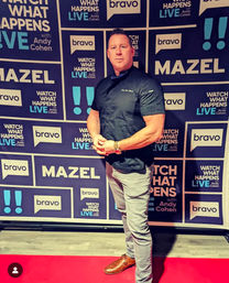 Man in a black chef jacket and gray pants striking a confident pose on a red carpet in front of a branded media wall at a TV studio event.