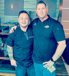 Two smiling chefs in black jackets pose with arms around each other inside a modern restaurant dining room with pendant lights and neatly set tables.