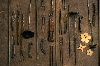 Flat-lay of blacksmith tools and hand-forged metalwork on a rustic workshop bench: twisted iron bars and hooks, hand-forged knives, a horseshoe and brass flower cutouts.
