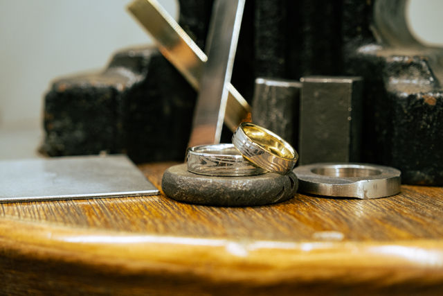 Close-up of two textured wedding bands—one gold-lined—resting on a jeweler's workbench among metal tools and rings on a wooden surface.