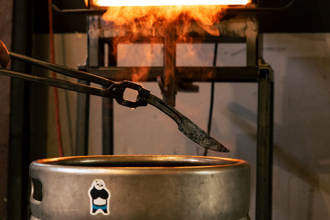 Blacksmith tongs holding a hot knife blade over an orange forge flame above a metal keg with a small cartoon sticker in an industrial workshop