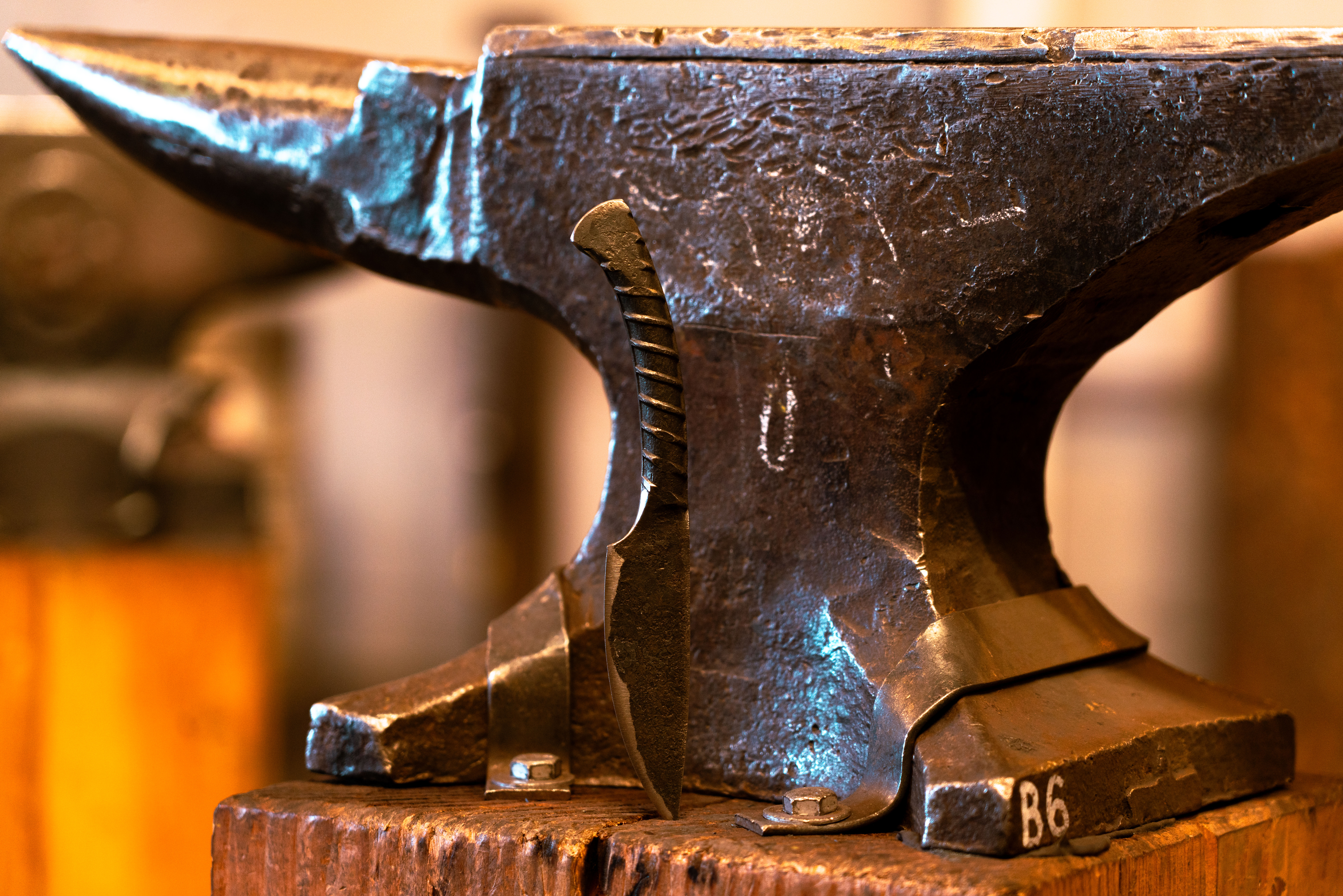 Close-up of a forged knife blade leaning against a weathered iron anvil on a wooden stump in a rustic blacksmith workshop