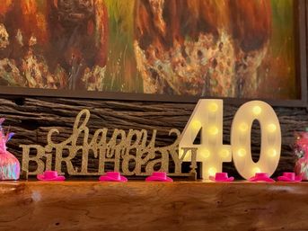 Festive 40th birthday mantel display with glittery gold "happy birthday" sign, illuminated marquee "40" numbers and small pink mini top hats on a wooden shelf against colorful abstract wall art.