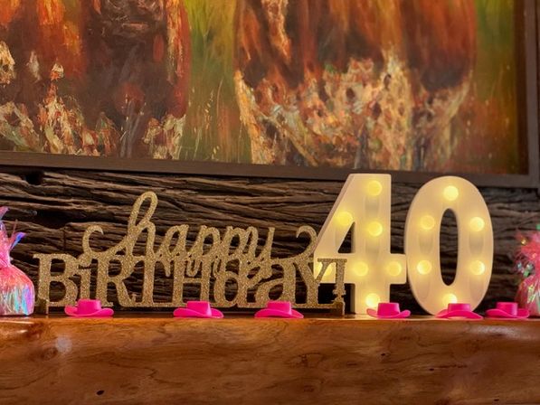 Festive 40th birthday mantel display with glittery gold "happy birthday" sign, illuminated marquee "40" numbers and small pink mini top hats on a wooden shelf against colorful abstract wall art.