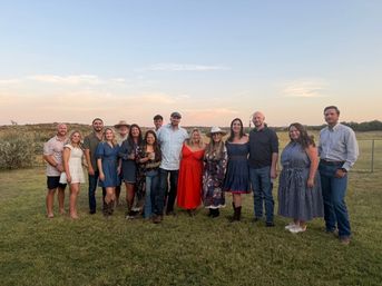 About 15 adults smiling and posing in a row on a grassy field at sunset, wearing casual and country-style outfits like dresses, hats, boots and jeans — outdoor group photo.