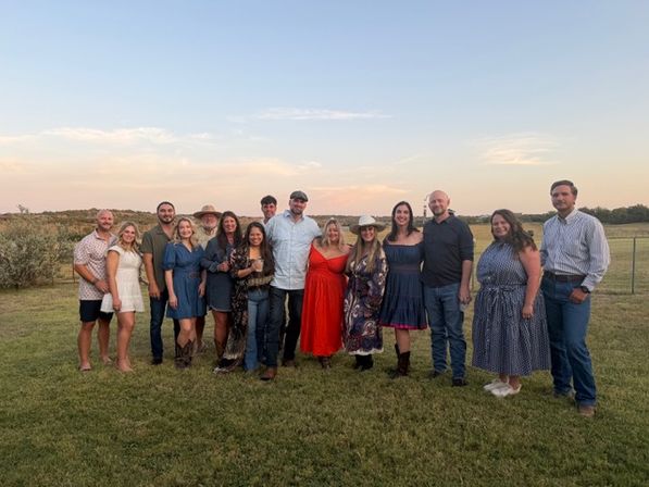 About 15 adults smiling and posing in a row on a grassy field at sunset, wearing casual and country-style outfits like dresses, hats, boots and jeans — outdoor group photo.
