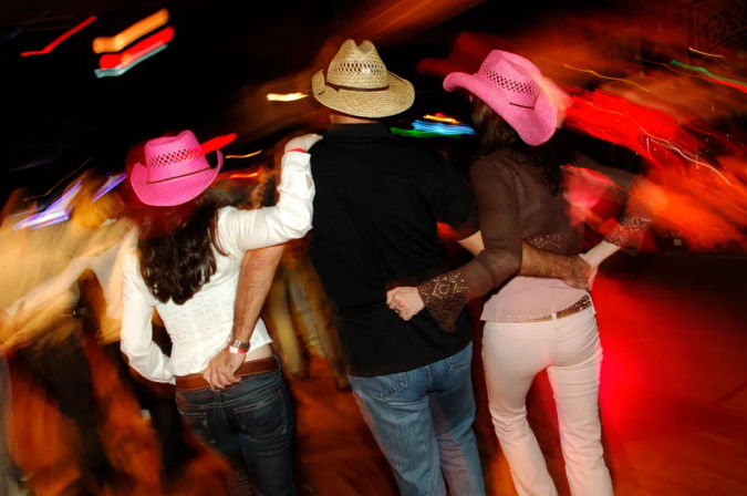 Three people arm-in-arm on a lively country-western dance floor at night — a straw cowboy hat between two pink cowboy hats, motion-blurred colorful lights and energetic crowd.