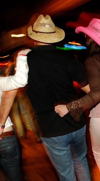 Three people arm-in-arm on a lively country-western dance floor at night — a straw cowboy hat between two pink cowboy hats, motion-blurred colorful lights and energetic crowd.