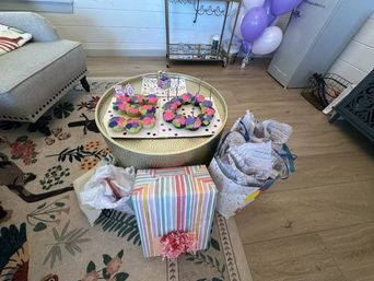 Home living room birthday setup: round coffee table with colorful frosted doughnuts and a '50' cake topper, striped wrapped gift with pink bow, gift bags, purple balloons, floral rug and hardwood floor.