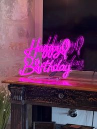 Cheerful pink neon 'Happy Birthday' acrylic sign glowing on a rustic wooden console table by an exposed brick wall, reflecting on a nearby TV — birthday party decor.
