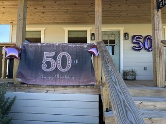 Front porch with wooden steps and railing decorated for a 50th birthday: purple glitter banner reading 50th Happy Birthday, matching star balloons and large 50 foil balloons on the house siding.
