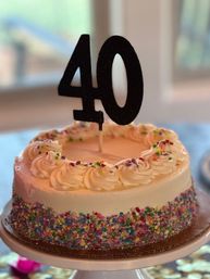 Round white frosted birthday cake with piped rosettes, colorful sprinkles around the sides, and a black "40" topper on a cake stand by a window