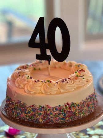 Round white frosted birthday cake with piped rosettes, colorful sprinkles around the sides, and a black "40" topper on a cake stand by a window