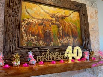 Rustic mantel beneath a large framed longhorn painting on a stone wall, styled as a cowboy-themed 40th birthday display with illuminated 40, pink mini cowboy hats and wrapped party favors.