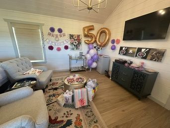 Cozy bright living room decorated for a 50th birthday with large gold '50' balloons, purple and white balloon cluster, 'Happy Birthday' banner, wrapped gifts on a floral rug, tufted sofa, bar cart and wall-mounted TV.