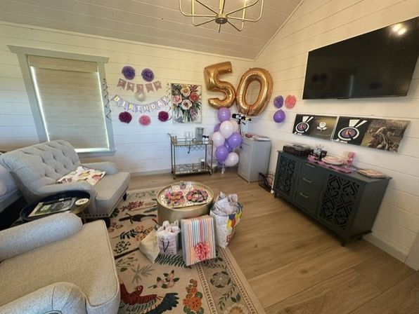 Cozy bright living room decorated for a 50th birthday with large gold '50' balloons, purple and white balloon cluster, 'Happy Birthday' banner, wrapped gifts on a floral rug, tufted sofa, bar cart and wall-mounted TV.