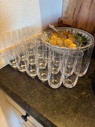 Rows of tall clear drinking glasses neatly lined up on a dark kitchen countertop beside a decorative ceramic bowl of lemons and limes and a wooden cutting board.