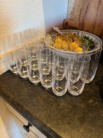 Rows of tall clear drinking glasses neatly lined up on a dark kitchen countertop beside a decorative ceramic bowl of lemons and limes and a wooden cutting board.