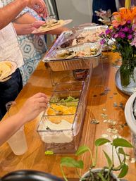 Indoor taco-bar buffet on a wooden table with aluminum trays of sliced meat and warm tortillas, clear bins of sour cream, shredded cheese, guacamole and toppings as guests assemble tacos next to a vase of flowers.